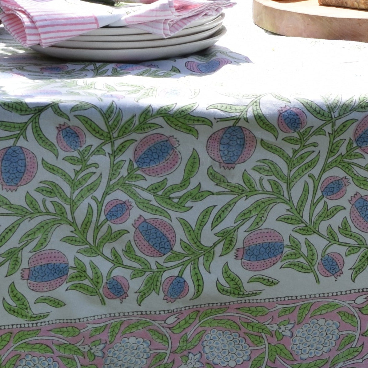 Hand Block Print Cotton Tablecloth – Perfectly Pomegranate close detail showing the all-over pink, blue and green pomegranate and trailing leaf block print repeat with decorative floral border, draped over a garden table in bright sunlight with stacked white plates and striped napkins in the background